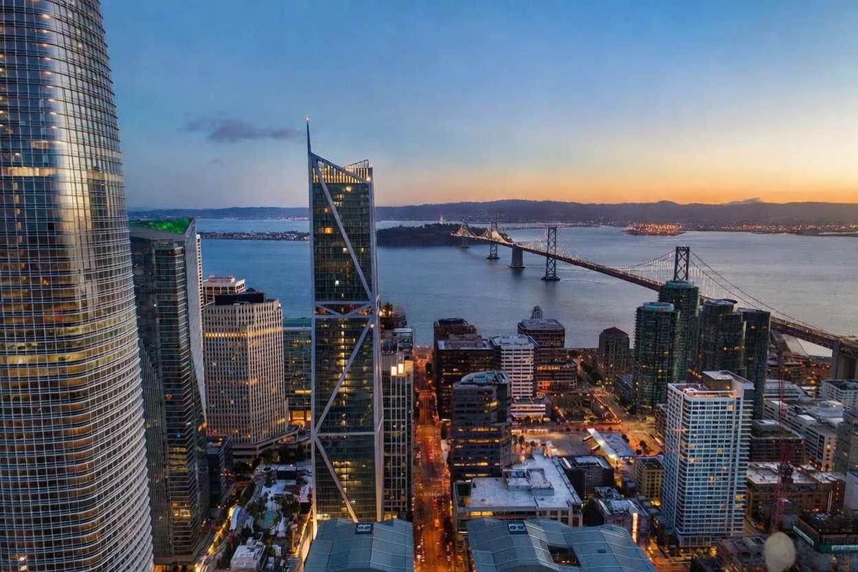 This high-altitude aerial view captures a stunning sunset over the San Francisco skyline, prominently featuring the Salesforce Tower and the iconic Bay Bridge in the background. The composition highlights the dense urban architecture, including modern skyscrapers with glass facades and the bustling city streets below. The golden hour lighting casts a warm glow across the bay and the city, creating a sophisticated and dramatic urban atmosphere.