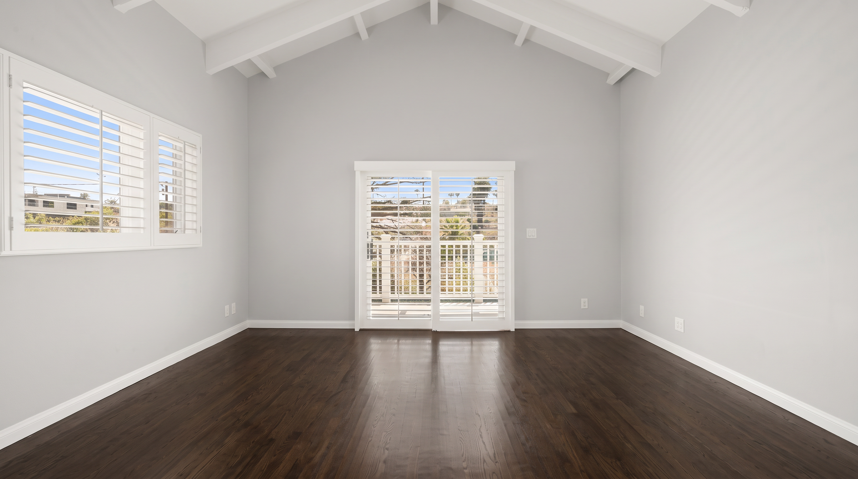 This spacious, unfurnished room features a high vaulted ceiling with exposed beams and light gray walls. The space is illuminated by natural light streaming through a large window on the left and a set of sliding glass doors that open to an outdoor area. The floor is currently unfinished plywood, providing a blank canvas for new flooring installation.