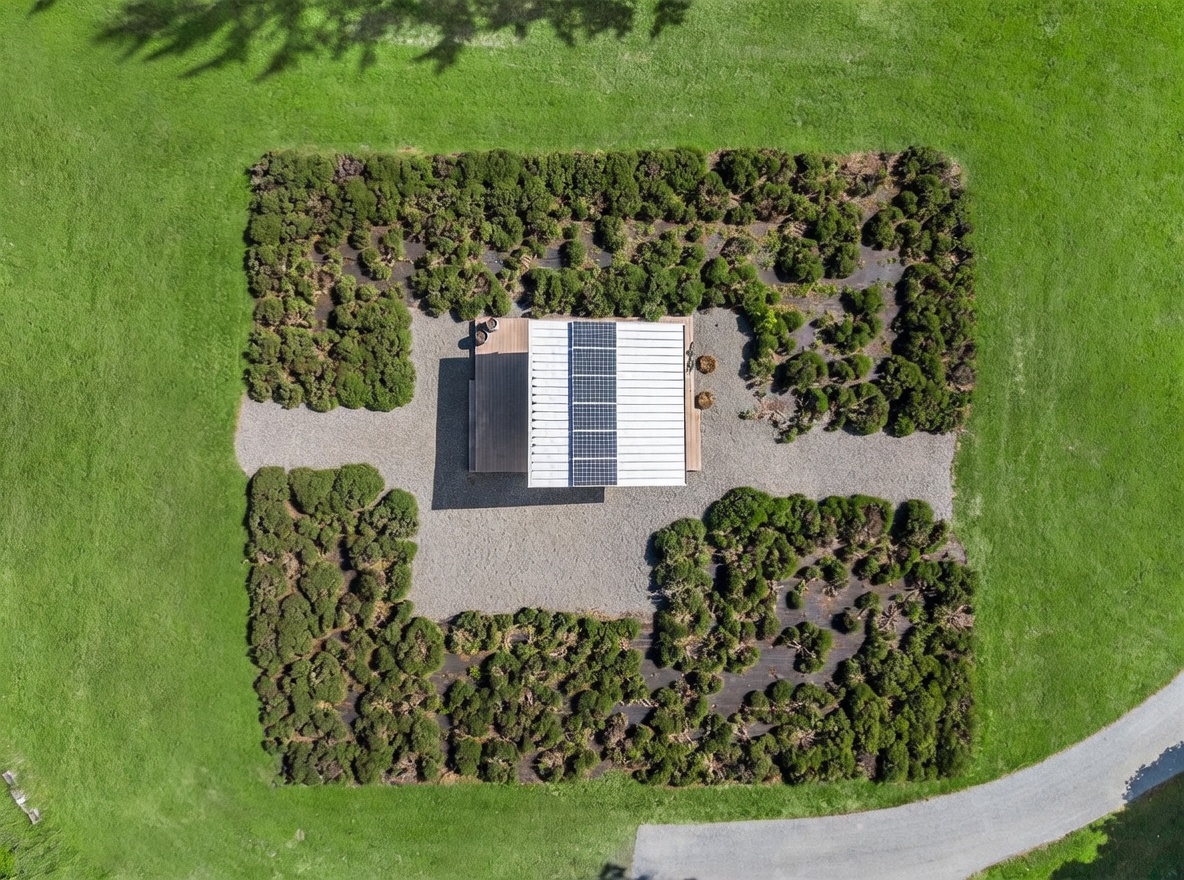 This high-angle aerial view captures a modern, flat-roofed structure centered within a manicured garden plot. The building features a prominent array of solar panels on its roof and is surrounded by a gravel perimeter, which is framed by dense, low-lying shrubbery set against a vast expanse of green lawn. A paved driveway curves into the bottom right corner, providing a clear perspective of the property's isolated and tranquil setting.