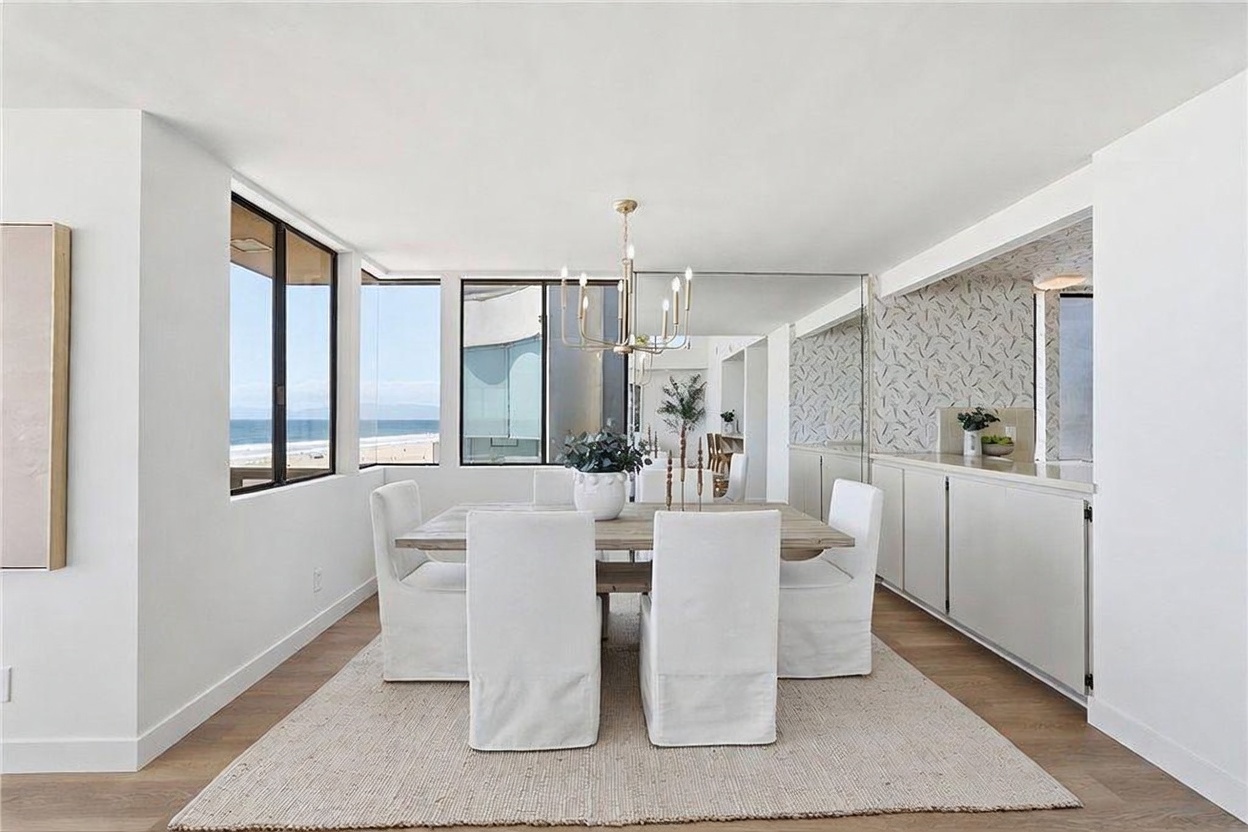This bright and airy dining room features a light wood table surrounded by four white slipcovered chairs, set atop a textured area rug. A modern chandelier hangs above the table, while a large mirror on the wall reflects the space and enhances the natural light streaming in from the large windows overlooking the ocean. To the right, a built-in sideboard with patterned wallpaper above provides additional storage and display space, creating a clean, coastal-inspired aesthetic.