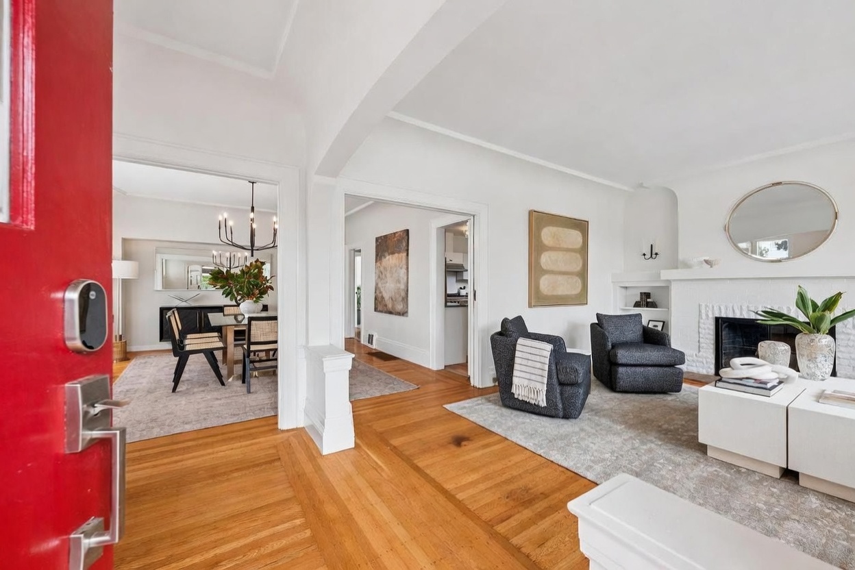 This bright and inviting living room features warm wood flooring and a neutral color palette, centered around a white fireplace with a round mirror above. Two dark gray armchairs provide comfortable seating, while an open floor plan allows for a clear view into the adjacent dining area. The perspective is captured from the entryway, emphasizing the home's spacious and airy flow.