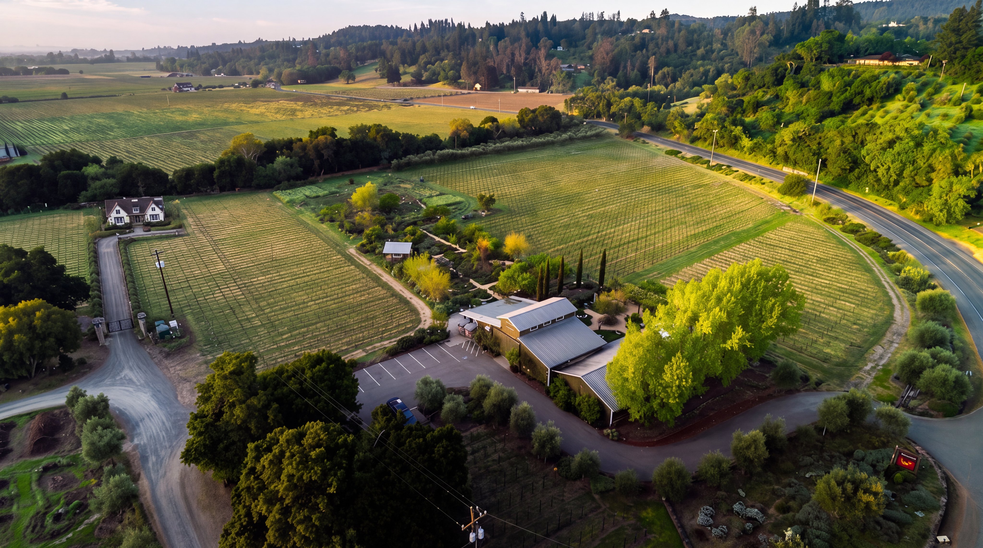 This stunning aerial view captures a sprawling vineyard estate featuring a modern barn-style building with a metal roof and a spacious parking lot. The property is surrounded by lush rows of vines, mature trees, and landscaped gardens, creating a serene and picturesque pastoral setting. A charming white farmhouse is also visible on the edge of the property, adding to the estate's overall rustic yet sophisticated appeal.