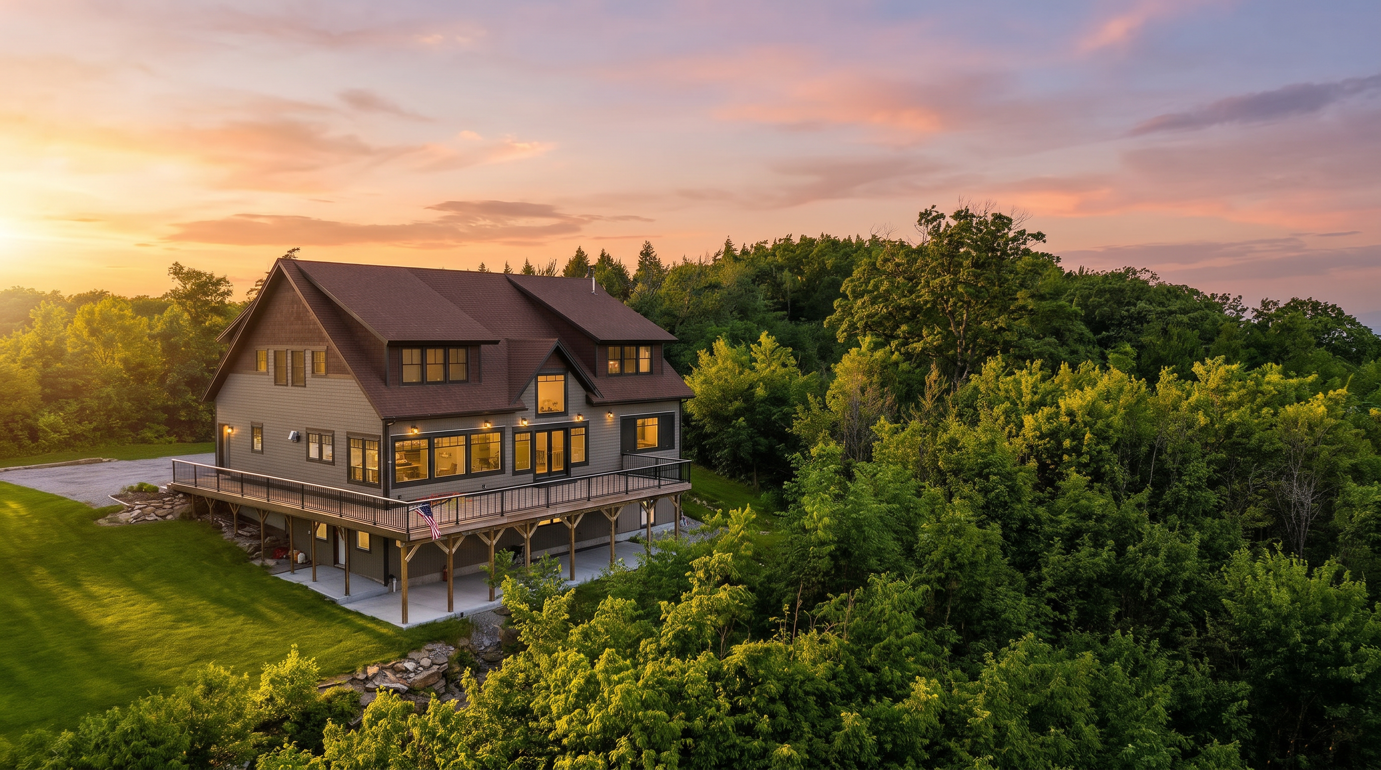 This elevated aerial perspective captures a multi-story home nestled against a dense, lush forest. The house features a large, elevated wooden deck that spans the rear, providing an expansive outdoor living space overlooking the surrounding greenery. The structure is finished in neutral-toned siding with a dark shingled roof, presenting a serene and private retreat set within a natural landscape.