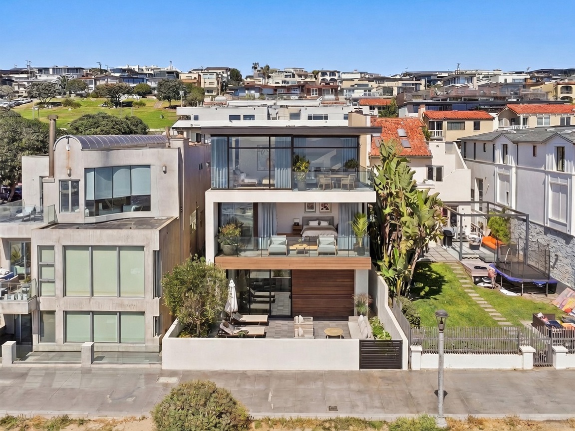 This contemporary three-story beachfront residence features a modern architectural design with expansive glass walls and balconies on the upper levels. The ground floor includes a private patio area with lounge seating, while the upper levels showcase open-concept living spaces and a bedroom with direct ocean views. The property is situated along a paved walkway, adjacent to a grassy yard and mature landscaping, offering a sophisticated coastal aesthetic.