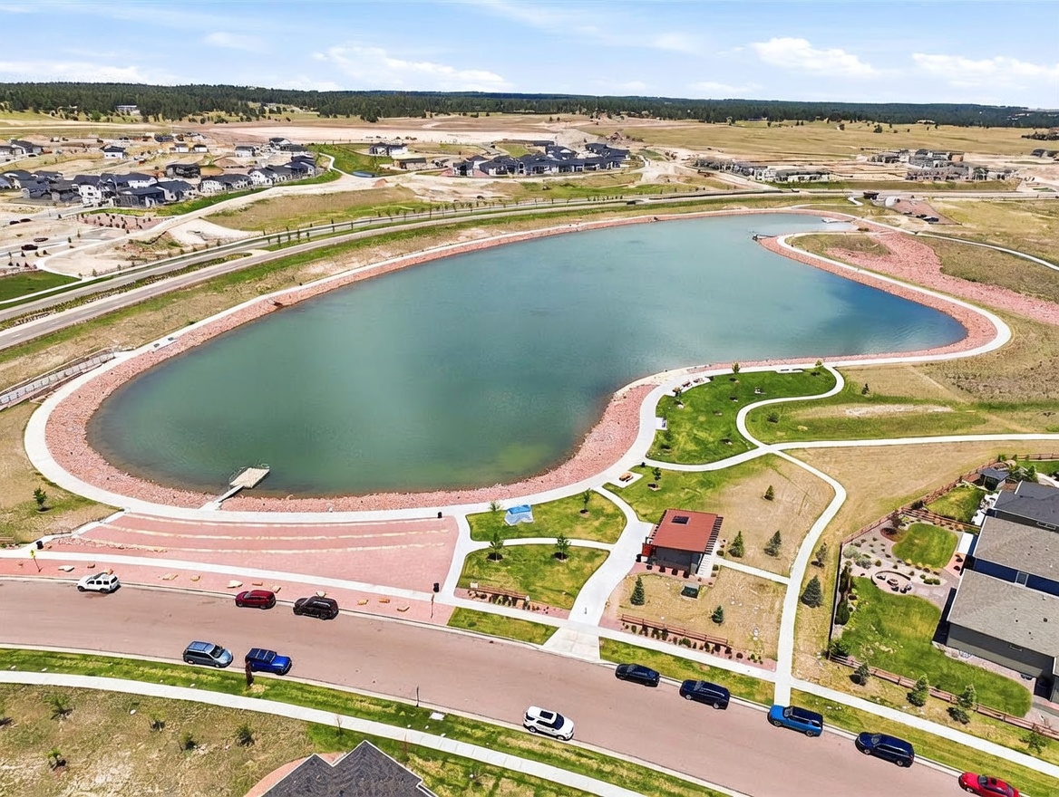 This high-angle aerial view captures a serene community lake surrounded by paved walking paths, manicured green spaces, and a small park structure. The perspective highlights the neighborhood's layout, showing residential streets with parked cars and a backdrop of expansive, undeveloped land. The scene conveys a peaceful, master-planned suburban atmosphere with convenient access to outdoor recreational amenities.