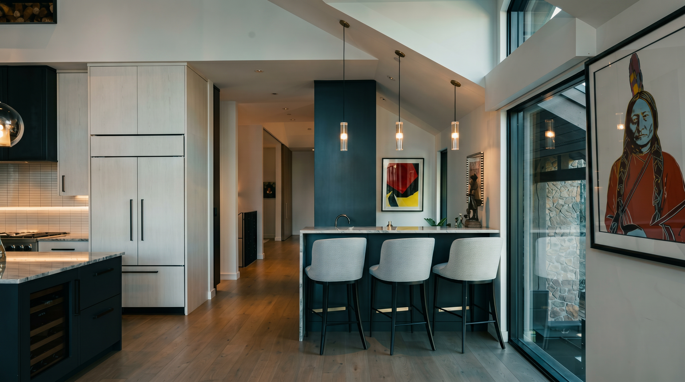 This modern kitchen features a sleek island with three white bar stools, set against a dark accent wall that anchors the space. The area is illuminated by three minimalist pendant lights and benefits from natural light streaming through large windows on the right. Light wood flooring flows throughout the open-concept layout, which includes white cabinetry and a wine cooler integrated into the island base.