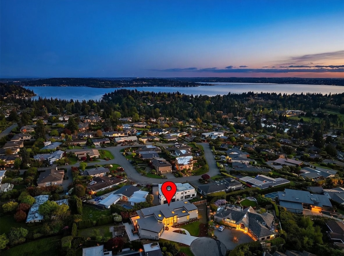 This high-angle aerial shot captures a modern, multi-level residential property at dusk, highlighted by a red location pin. The home features a contemporary architectural design with flat rooflines, warm exterior lighting, and a paved driveway, set within a lush, suburban neighborhood overlooking a large body of water. The cinematic perspective emphasizes the home's prominent position within the community and its scenic surroundings.
