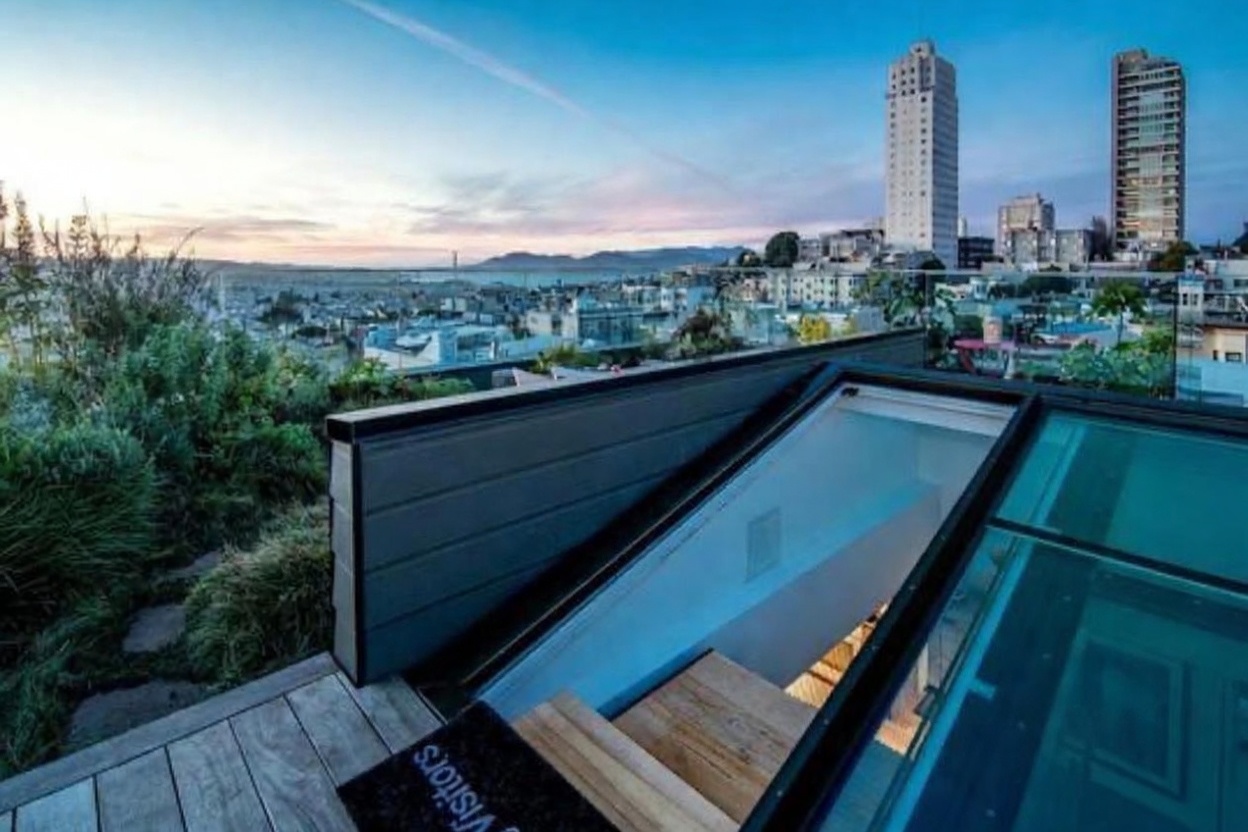 This elevated deck offers a stunning panoramic view of the city skyline at dusk, featuring a modern glass-enclosed staircase leading down into the home. The deck is constructed with light-toned wood planks and includes a 'Visitors' mat near the entrance. The perspective is from a high vantage point, emphasizing the seamless transition between the outdoor living space and the interior architecture.