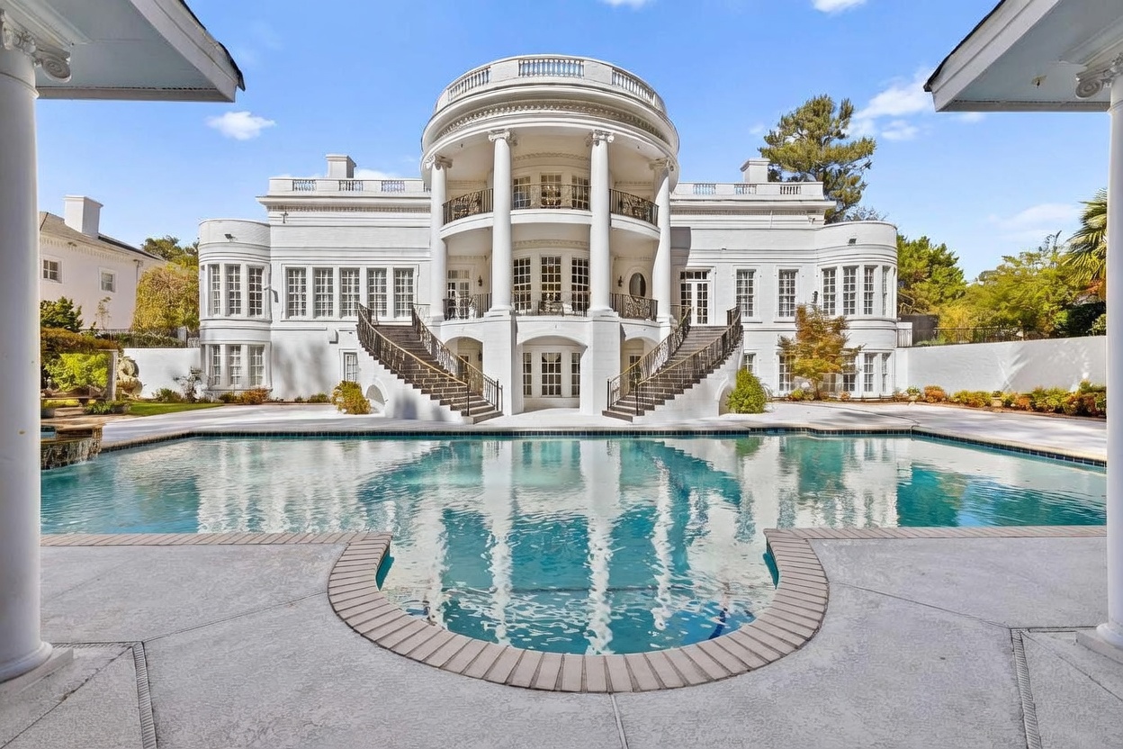 This grand, neoclassical-style mansion features a symmetrical white facade with a prominent central rotunda and dual sweeping staircases leading to the main level. A large, custom-shaped swimming pool occupies the foreground, reflecting the architectural details of the home under a clear blue sky. The property exudes luxury and elegance, framed by manicured landscaping and white perimeter walls.