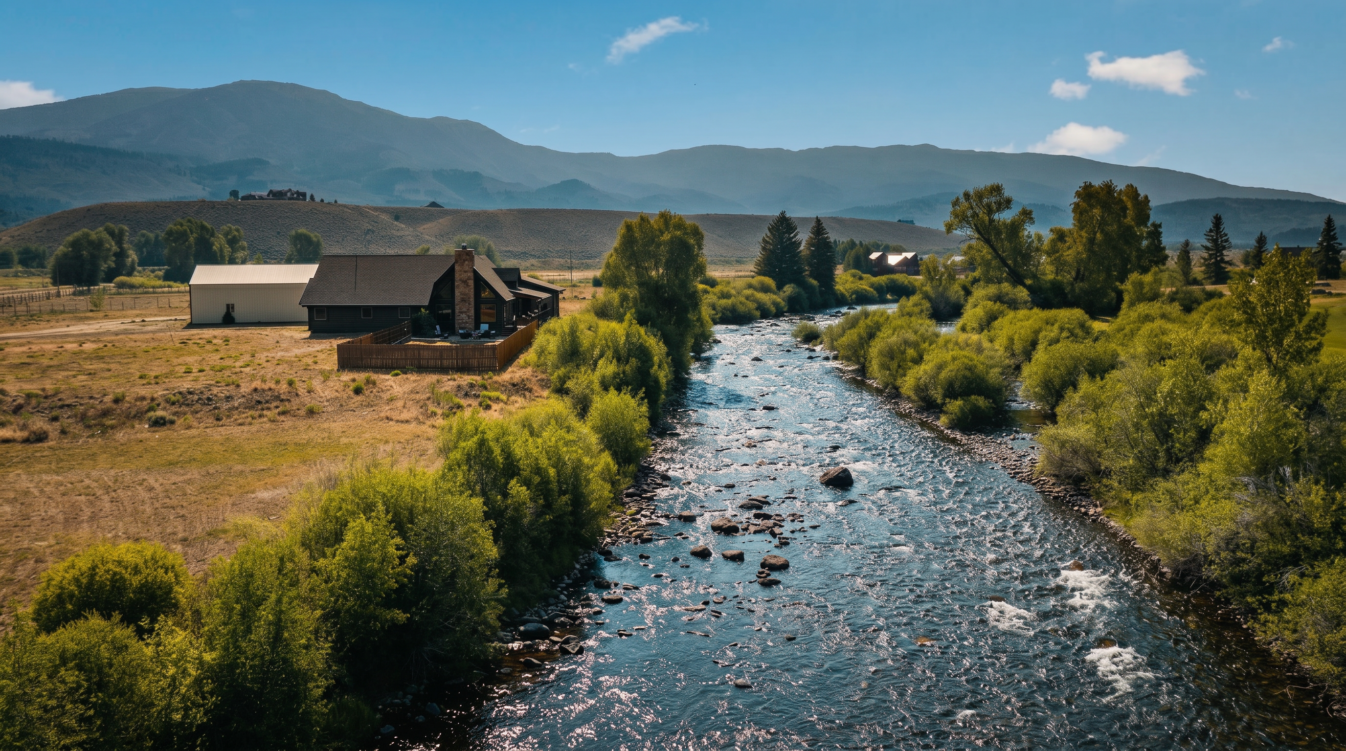 This aerial perspective captures a rustic-style home situated along the banks of a flowing river, framed by lush green vegetation and rolling hills in the distance. The property includes a large, light-colored outbuilding and a fenced-in yard area, emphasizing its secluded, natural setting. The bright, sunny sky and expansive landscape highlight the tranquil and scenic nature of this rural estate.