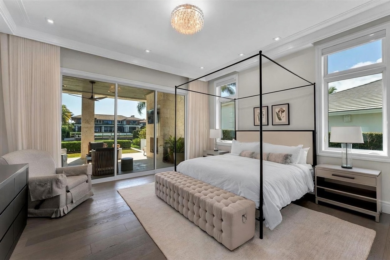 This spacious primary bedroom features a modern four-poster bed as the centerpiece, complemented by a tufted upholstered bench at the foot. Large glass sliding doors provide a seamless transition to a covered outdoor patio, while light-toned walls and wood flooring create an airy, sophisticated atmosphere. The room is accented by a crystal chandelier and symmetrical bedside lamps, offering a refined and tranquil living space.