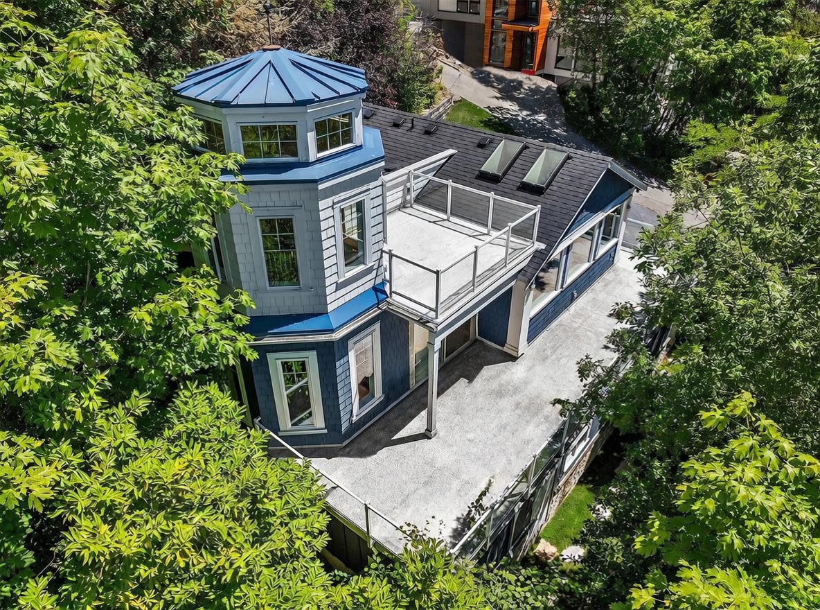 This high-angle aerial shot captures a unique, multi-level home featuring a prominent blue turret and a spacious rooftop deck with glass railings. The house is nestled among lush green trees, showcasing a modern architectural style with shingle siding and a dark shingled roof. The perspective highlights the property's integration with its wooded surroundings and the expansive outdoor living spaces.