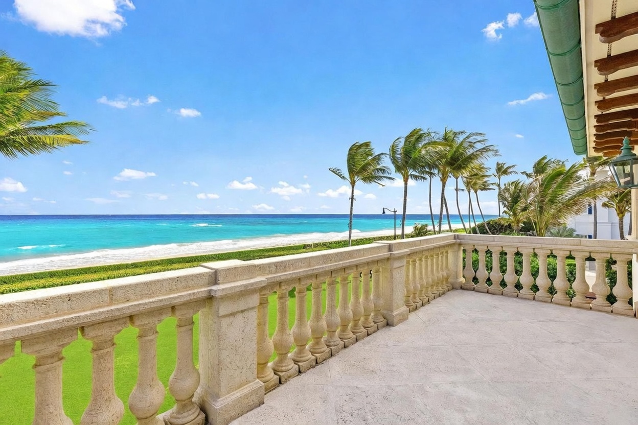 This elevated balcony features a classic stone balustrade and light-toned stone flooring, offering an expansive, unobstructed view of the turquoise ocean and sandy beach. The scene is framed by lush green lawn below and a row of swaying palm trees, creating a serene and luxurious coastal atmosphere. The perspective captures the seamless transition between the private outdoor living space and the natural beauty of the shoreline.