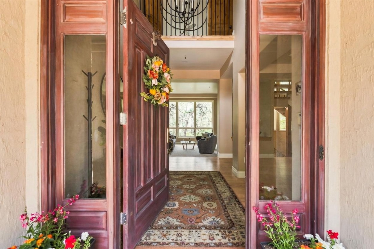 This perspective captures the transition from a covered porch into a grand foyer through a set of ornate, dark-stained wooden double doors adorned with a floral wreath. The entryway features a patterned area rug leading into a bright, open-concept interior with a view of a living area and large windows in the distance. A decorative chandelier hangs above the foyer, and potted flowers frame the threshold, creating a welcoming and elegant first impression.