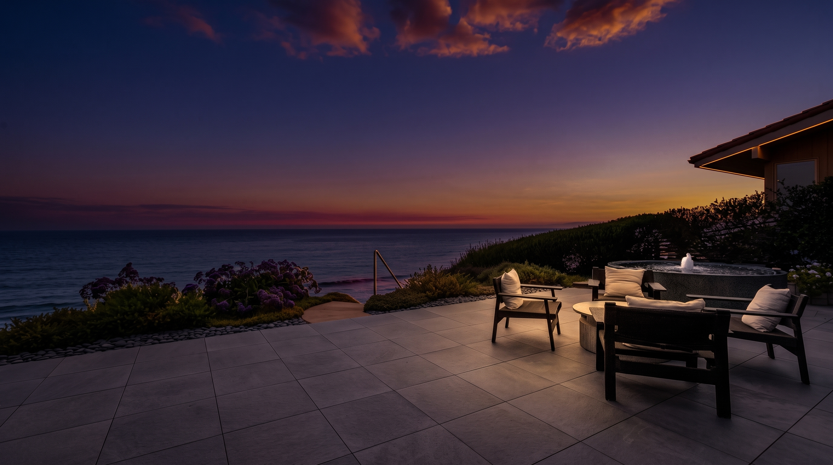 This expansive oceanfront patio features large, light-toned stone pavers and a comfortable seating area arranged around a circular fire pit. The space is framed by lush coastal landscaping and a circular spa, all set against a panoramic, unobstructed view of the sea under a bright, cloudy sky. The perspective captures the seamless transition between the refined outdoor living space and the natural beauty of the coastline.