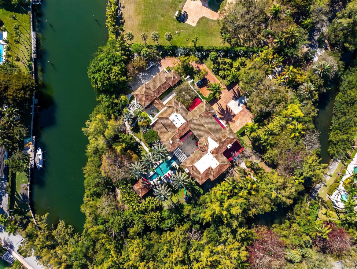 This high-angle aerial view captures a sprawling, Mediterranean-style estate nestled along a lush, tree-lined canal. The property features a complex roofline with terracotta tiles, a central courtyard, and a private swimming pool area surrounded by tropical landscaping. A large boat is docked along the canal, emphasizing the waterfront lifestyle and the secluded, expansive nature of the grounds.