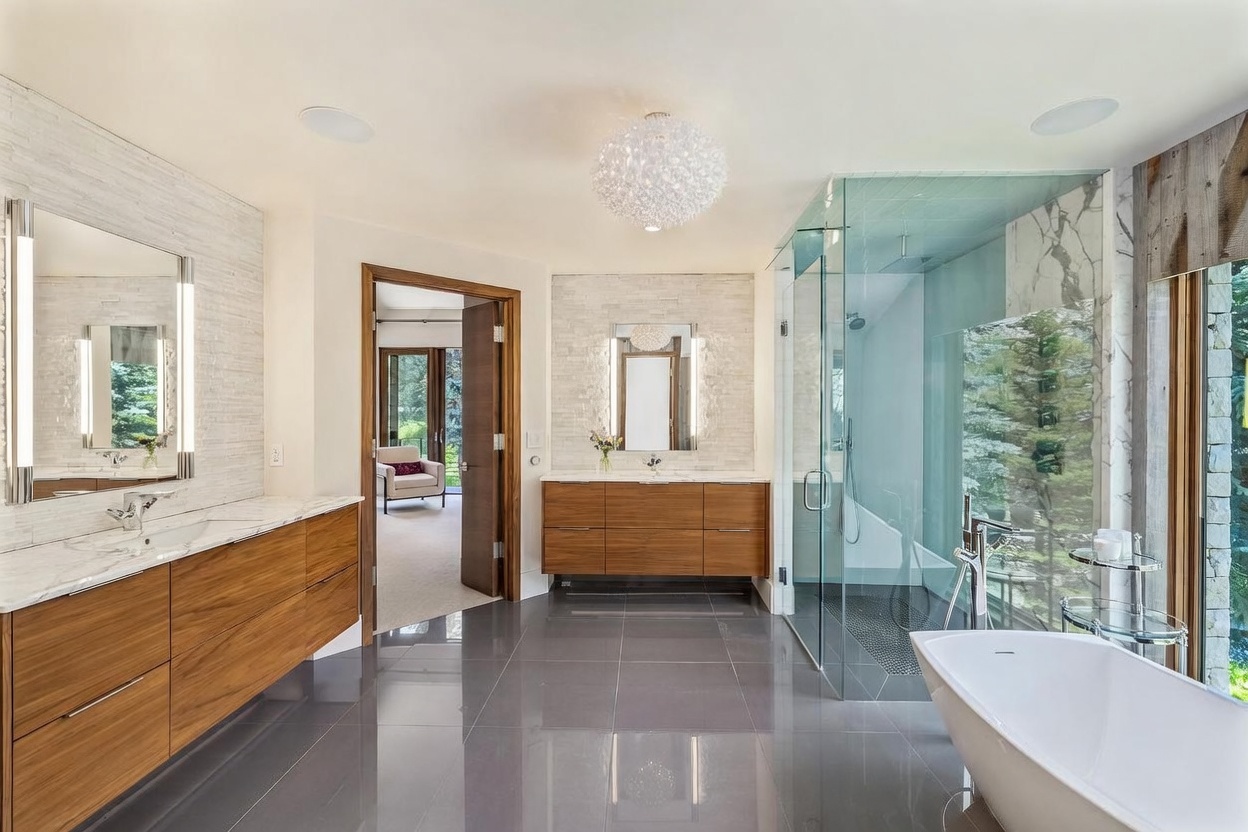 This luxurious primary bathroom features a double vanity with warm-toned wood cabinetry and stone countertops, complemented by a large glass-enclosed walk-in shower and a freestanding soaking tub. The space is finished with polished gray tile flooring and textured stone accent walls, while a large window provides a serene view of the surrounding forest. A modern, spherical chandelier serves as a focal point, creating an elegant and spa-like atmosphere.