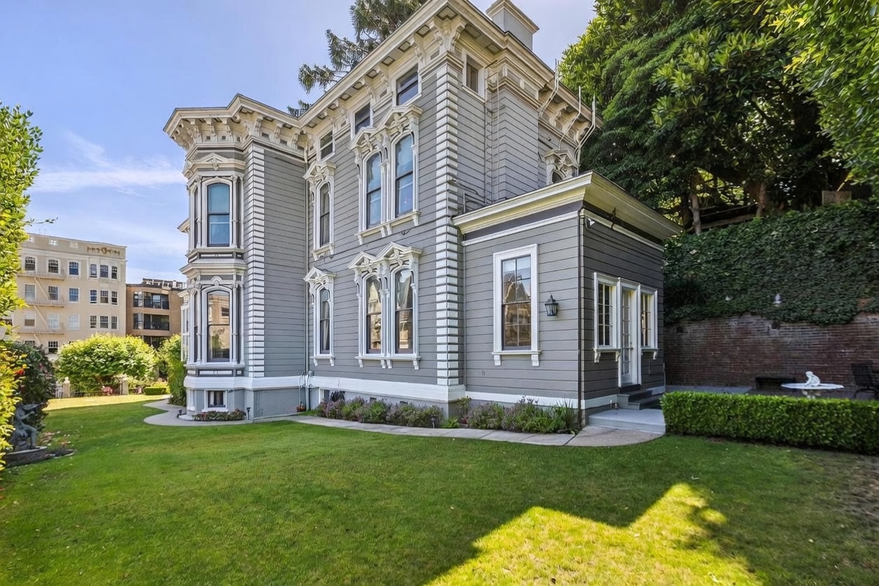 This image captures the side profile of a historic, multi-story Victorian-style home painted in a soft grey with crisp white trim. The architecture features a prominent bay window, intricate decorative molding, and a well-maintained lawn that leads to a stone-retaining wall covered in ivy. The perspective highlights the home's classic character and its integration into a lush, landscaped outdoor space.