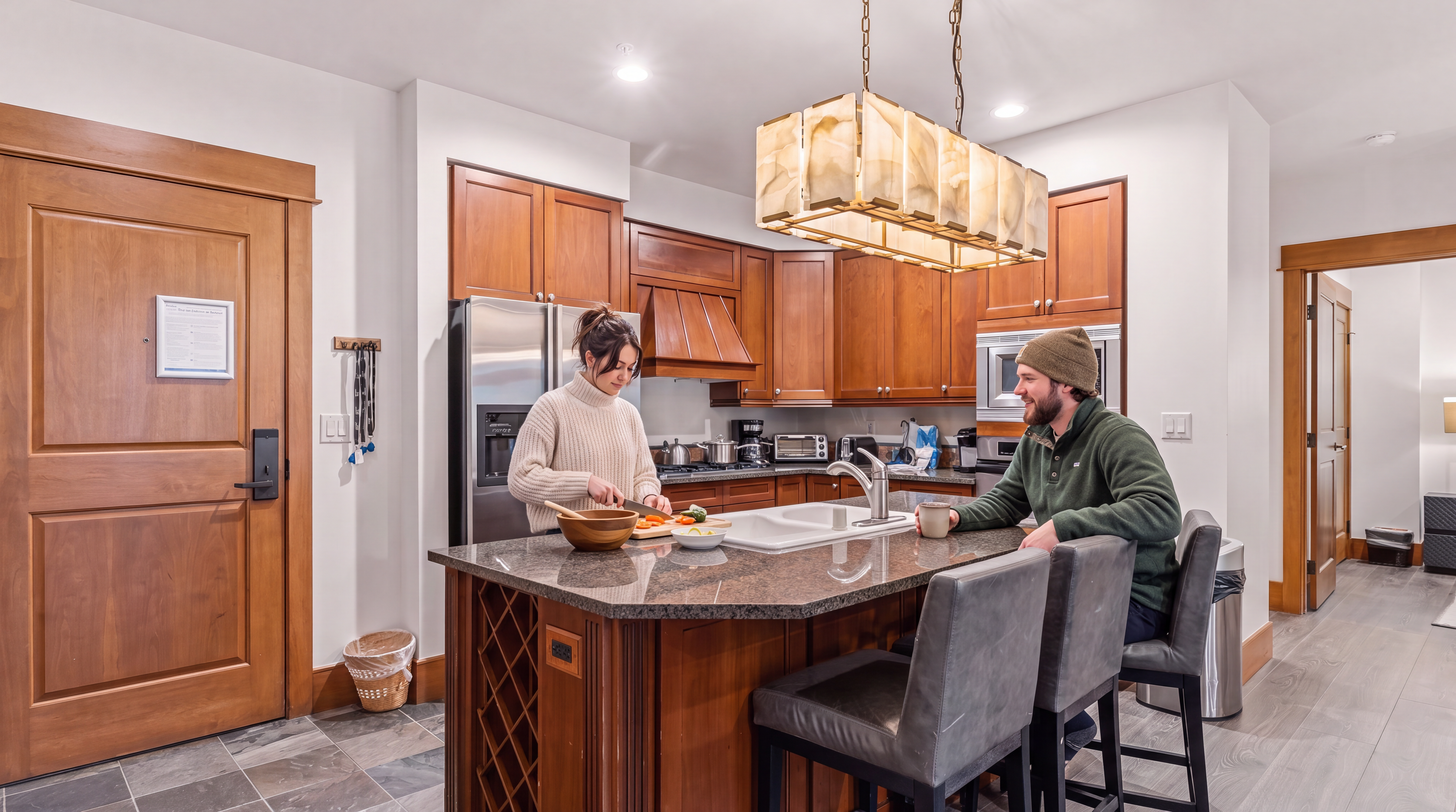 This kitchen features warm-toned wood cabinetry, a central island with a stone countertop, and stainless steel appliances. A distinctive rectangular pendant light fixture hangs above the island, which includes integrated wine storage and bar seating. The space is finished with a combination of stone-look tile flooring and wood-style flooring, creating a functional and inviting culinary area.