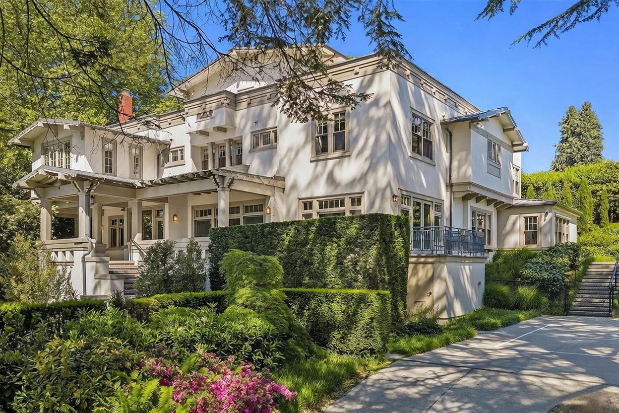 This elegant, multi-story residence features a classic architectural style with light-colored stucco walls and intricate trim details. The property is surrounded by lush, manicured landscaping, including tall hedges and vibrant flowering bushes, and includes a paved driveway leading toward the rear. The perspective captures the home's grand scale and sophisticated curb appeal under a clear blue sky.