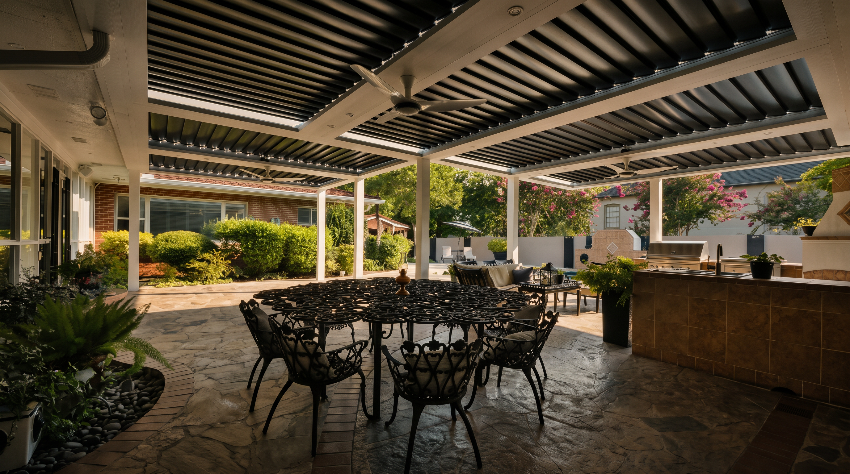 This spacious covered patio features a sophisticated louvered roof system, providing adjustable shade over a large, ornate metal dining set. The area includes a built-in outdoor kitchen with tiled counters and a grill, set against a backdrop of lush greenery and mature landscaping. The stone-paved flooring and ceiling fans create a comfortable, inviting atmosphere for outdoor entertaining.