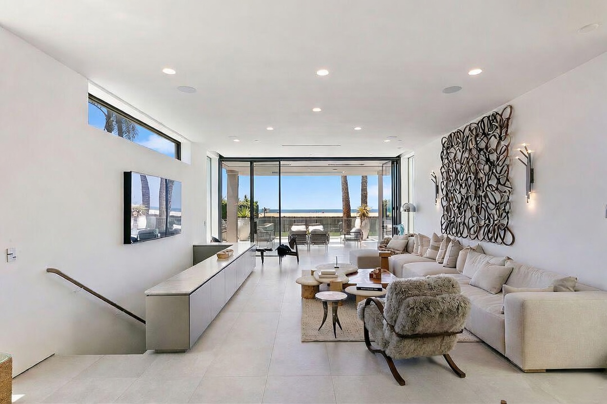 This bright and airy living room features a minimalist aesthetic with light-toned tile flooring and white walls. A large, textured wall sculpture serves as a focal point above a long, neutral-toned sectional sofa, while a plush, fur-covered armchair adds texture to the seating area. The space opens up through expansive glass doors to a patio with a direct view of the ocean, creating a seamless indoor-outdoor living experience.