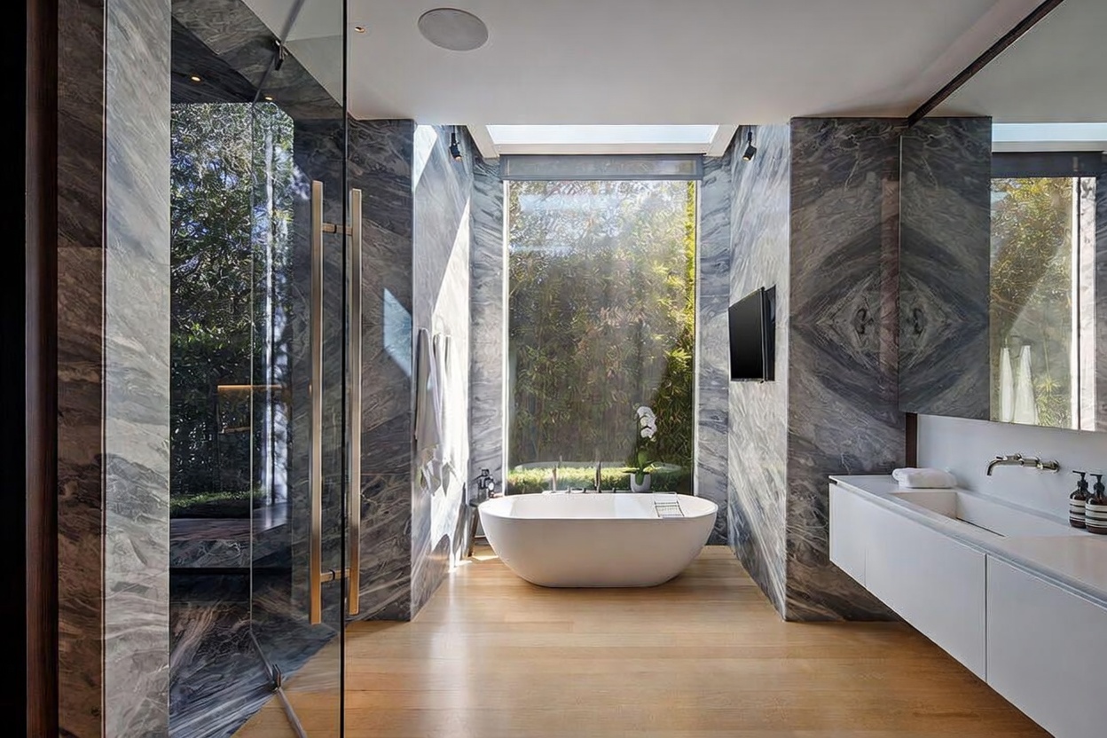 This luxurious primary bathroom features striking dark, veined stone walls that contrast with light wood flooring and a minimalist white vanity. A freestanding soaking tub is positioned in front of a large window that offers a serene view of lush greenery, while a glass-enclosed shower stall sits to the left. The space is illuminated by natural light and recessed ceiling fixtures, creating a sophisticated and tranquil spa-like atmosphere.
