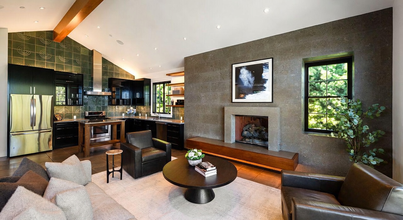 This open-concept living area seamlessly integrates a modern kitchen with a cozy lounge space, characterized by a striking stone-clad chimney breast and dark cabinetry. The room features a vaulted ceiling with exposed beams, a large round coffee table, and comfortable leather seating, all anchored by a neutral area rug. A large window provides natural light and a view of the greenery outside, creating a sophisticated and inviting atmosphere.