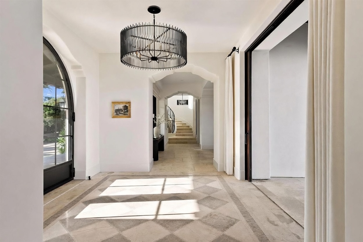 This elegant entryway features a sophisticated diamond-patterned stone floor and a striking, large black drum chandelier that serves as a focal point. The space transitions through a series of arched openings, leading the eye toward a staircase in the distance, while a large arched glass door on the left invites natural light into the foyer. The neutral color palette and clean architectural lines create a bright, airy, and welcoming atmosphere.