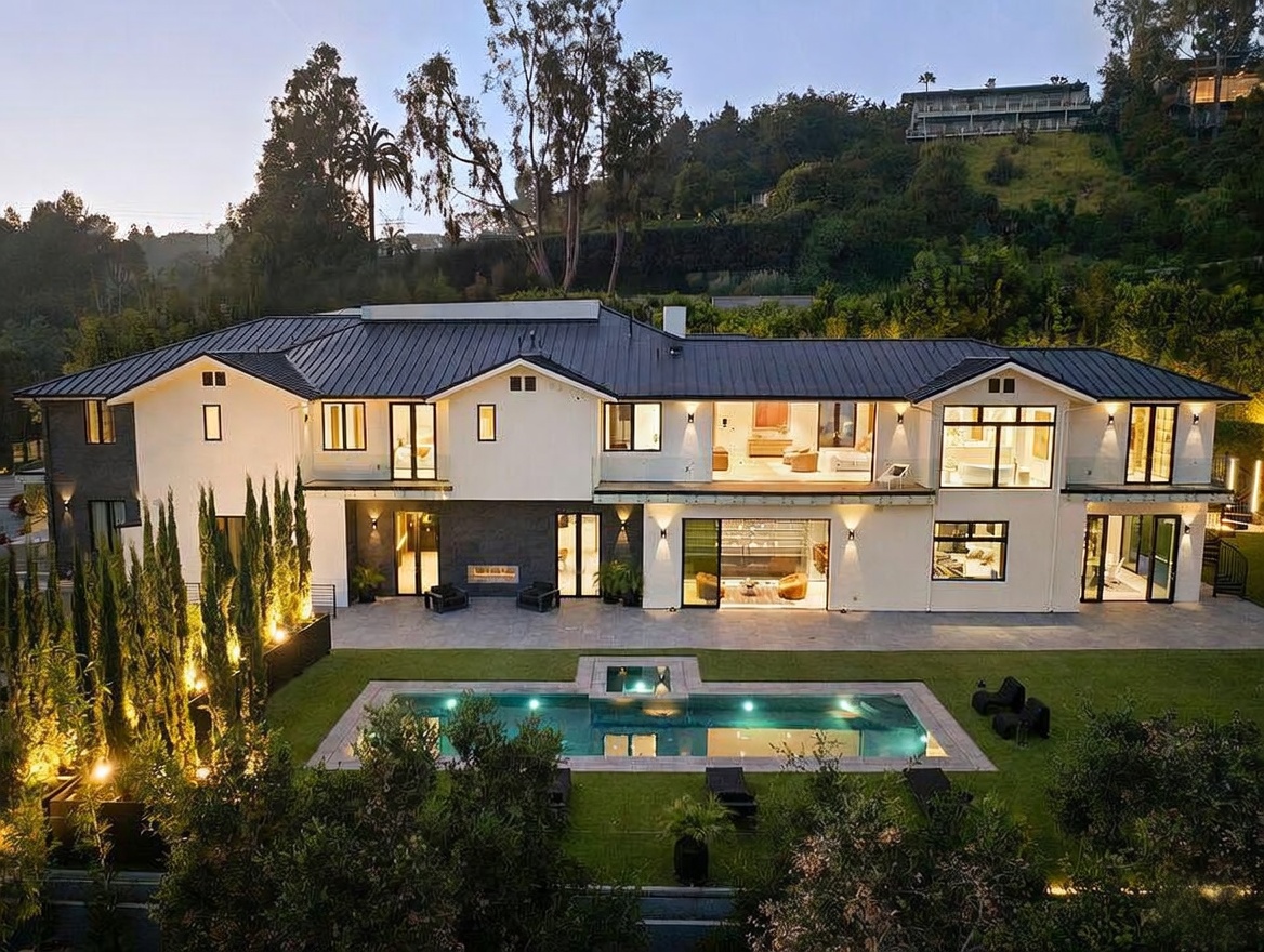 This expansive two-story luxury residence features a modern aesthetic with a dark metal roof, crisp white stucco walls, and dark stone accents. The rear facade opens onto a manicured lawn with a rectangular swimming pool and integrated spa, illuminated by warm landscape lighting. Large glass doors and windows create a seamless transition between the interior living spaces and the outdoor patio area.
