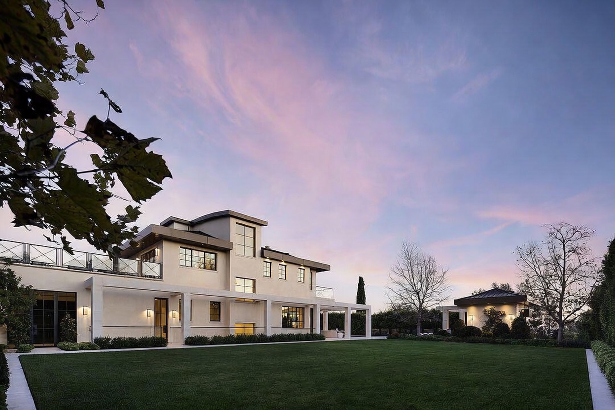 This rear view of a modern luxury residence showcases a sprawling, manicured lawn leading up to a multi-level structure with clean architectural lines and a neutral facade. The home features a covered patio area with square columns, large windows, and an upper-level terrace with metal railings. A smaller, matching detached structure sits to the right, all set against a soft, twilight sky.