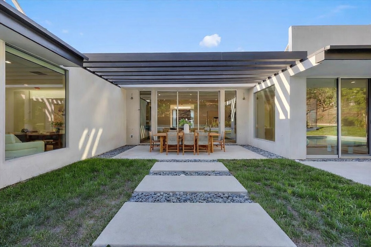 This modern outdoor living space features a covered patio area with a dining set, framed by a dark-toned pergola structure. A stone-paved walkway leads through a manicured lawn toward the patio, which is flanked by large glass sliding doors that offer a seamless transition to the interior. The clean lines and neutral color palette create a sophisticated, contemporary aesthetic perfect for outdoor entertaining.