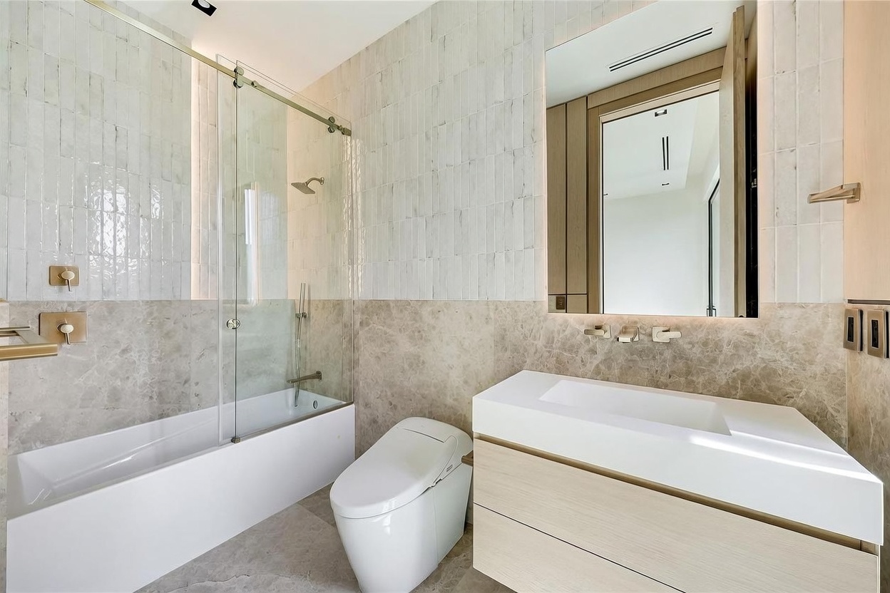 This modern bathroom features a sleek, wall-mounted vanity with a white countertop and light wood cabinetry. A glass-enclosed shower and tub combination is accented by neutral stone wall tiles and vertical textured wall cladding. The space is completed by a contemporary smart toilet and a large, frameless mirror, creating a bright and sophisticated atmosphere.