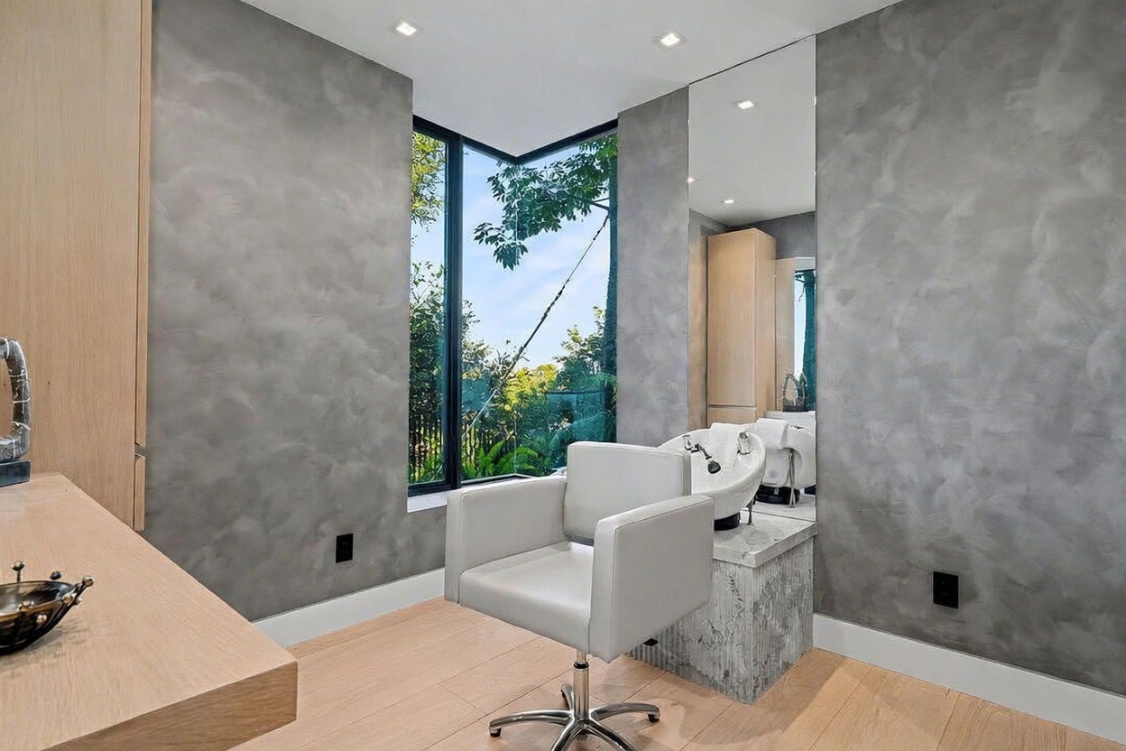 This image depicts a professional-style salon or spa room within a home, featuring a dedicated hair washing station with a sink and a swivel chair. The space is finished with textured gray walls, light wood flooring, and a large mirror that reflects the room and the outdoor greenery visible through the corner windows. The overall aesthetic is sleek and modern, designed for personal grooming and relaxation.