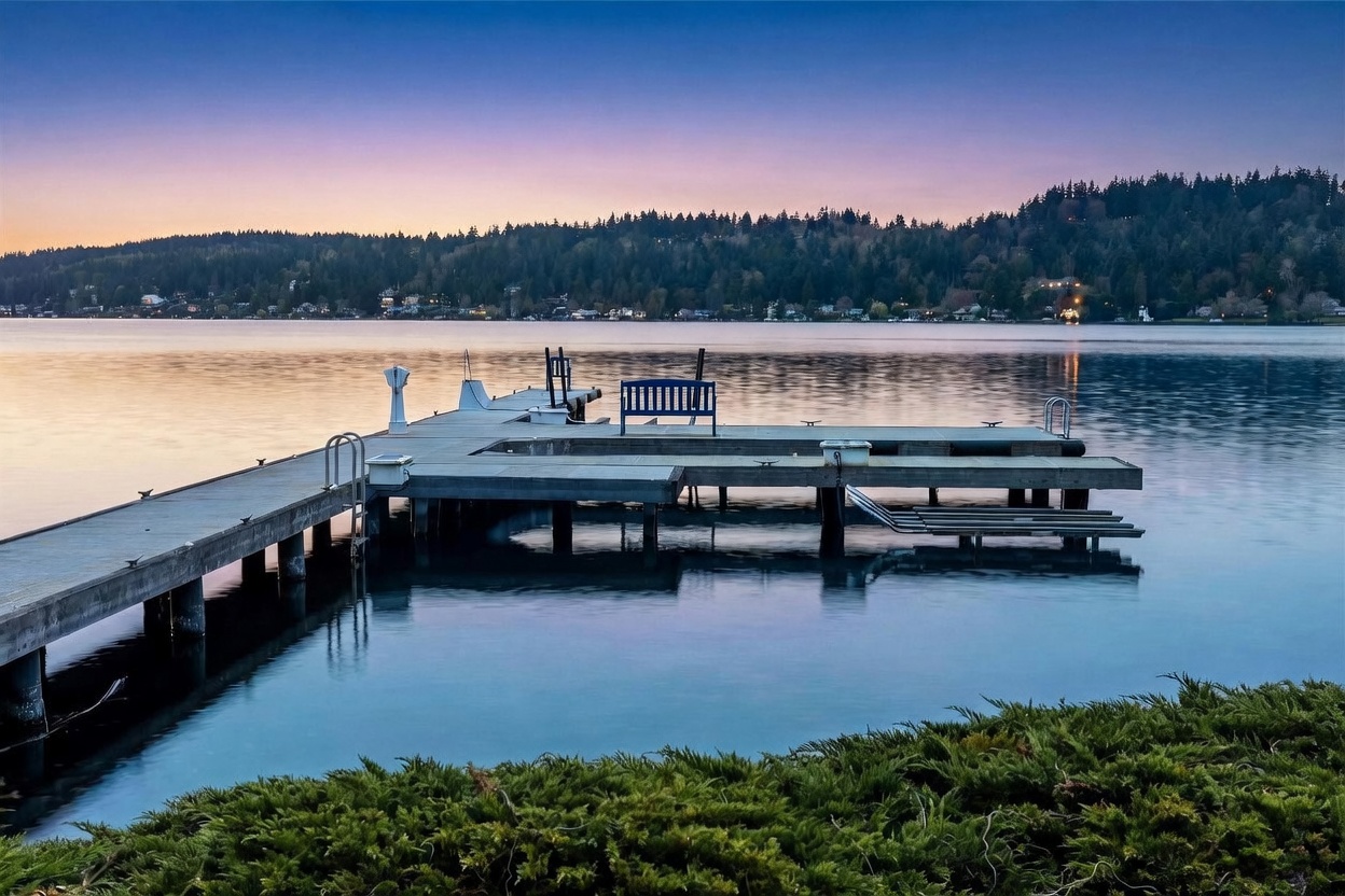 This serene waterfront view captures a private wooden dock extending into a calm lake during a soft, twilight sunset. The foreground features lush green shrubbery, while the background showcases a dense, tree-lined shoreline under a gradient purple and orange sky. The perspective is low and cinematic, emphasizing the peaceful atmosphere and the direct water access provided by the property.