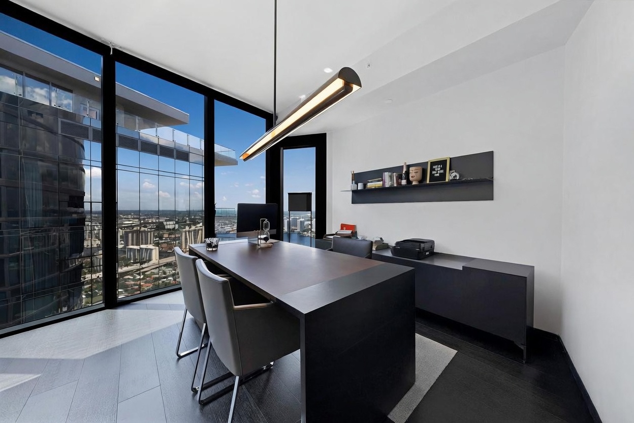 This modern home office features a sleek, dark-toned desk positioned to take advantage of the expansive, floor-to-ceiling windows that offer a panoramic city view. The room is minimalist in design, accented by a long, linear pendant light fixture and a wall-mounted shelf displaying decorative items. The dark wood flooring and monochromatic color palette create a sophisticated and professional atmosphere suitable for a high-end urban residence.