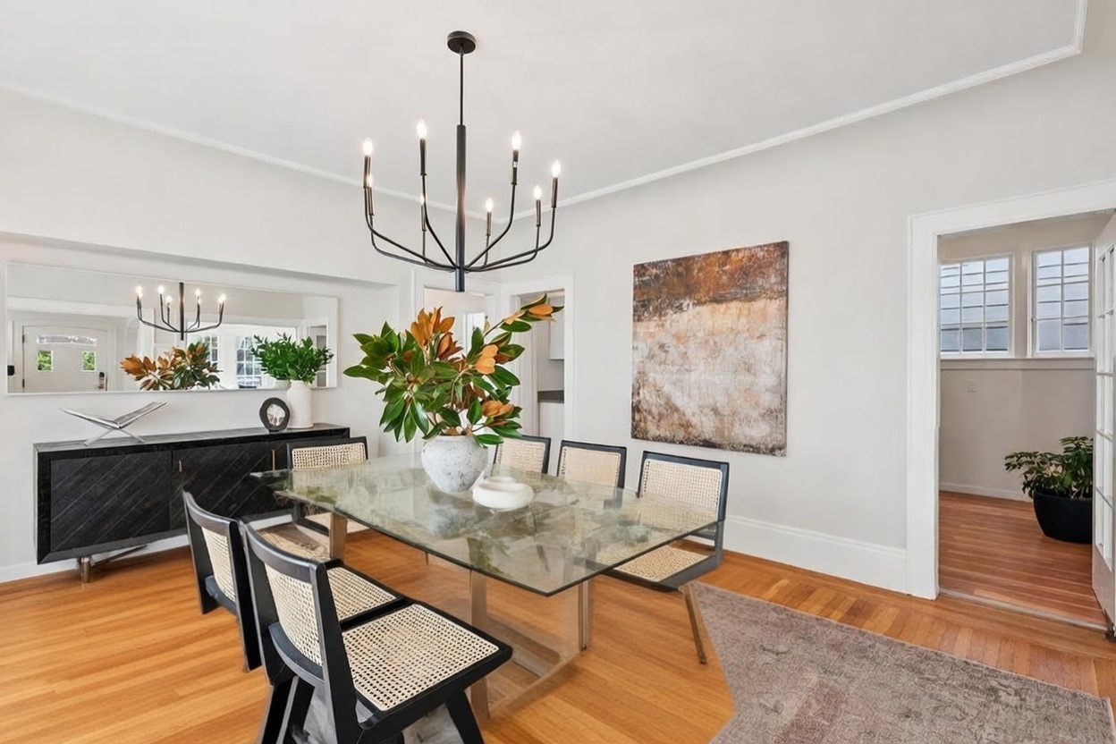 This elegant dining room features a glass-topped table surrounded by six black-framed chairs with woven cane seats. A striking black chandelier hangs above the table, which is centered on a large, neutral-toned area rug atop hardwood floors. The room is accented by a dark sideboard, a large abstract wall painting, and a mirror that reflects the entryway, creating a bright and sophisticated atmosphere.