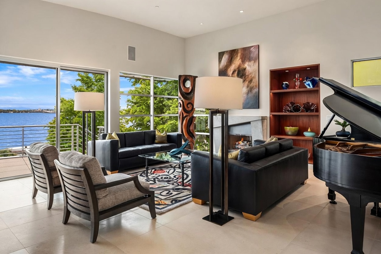 This sophisticated living room features a contemporary design with a grand piano, two black leather sofas, and a pair of gray upholstered armchairs arranged around a glass-topped coffee table. A tall, sculptural art piece stands beside a fireplace with a concrete-style surround, while a built-in wooden shelving unit displays decorative objects. Large windows provide an expansive, unobstructed view of the water, filling the space with natural light and creating a serene, upscale atmosphere.