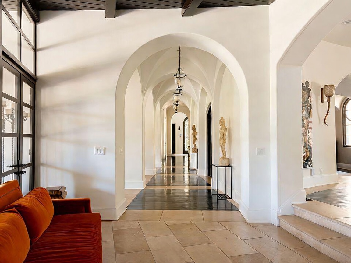 This elegant hallway features a series of dramatic white arches that create a sense of depth and architectural grandeur. The flooring transitions from large, light-toned stone tiles in the foreground to a dark, polished wood inlay that runs through the center of the arched corridor. An orange velvet sofa sits to the left, while minimalist pedestals displaying sculptures add a sophisticated, gallery-like aesthetic to the space.
