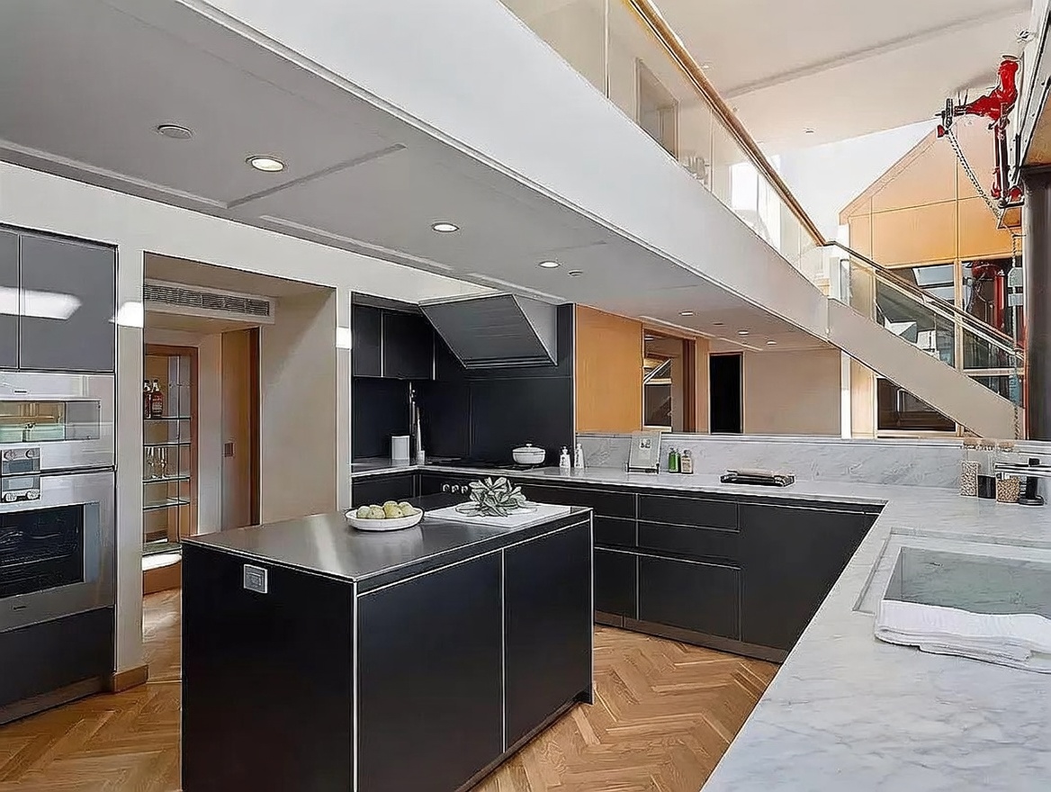 This modern, high-end kitchen features a sleek central island with a metallic finish and dark cabinetry, complemented by light stone countertops. The space is defined by an open-concept layout with a mezzanine level above, showcasing a glass railing and a striking red industrial-style pipe feature. The herringbone-patterned wood flooring adds warmth to the contemporary design, while the perspective captures the seamless flow between the cooking area and the architectural staircase.