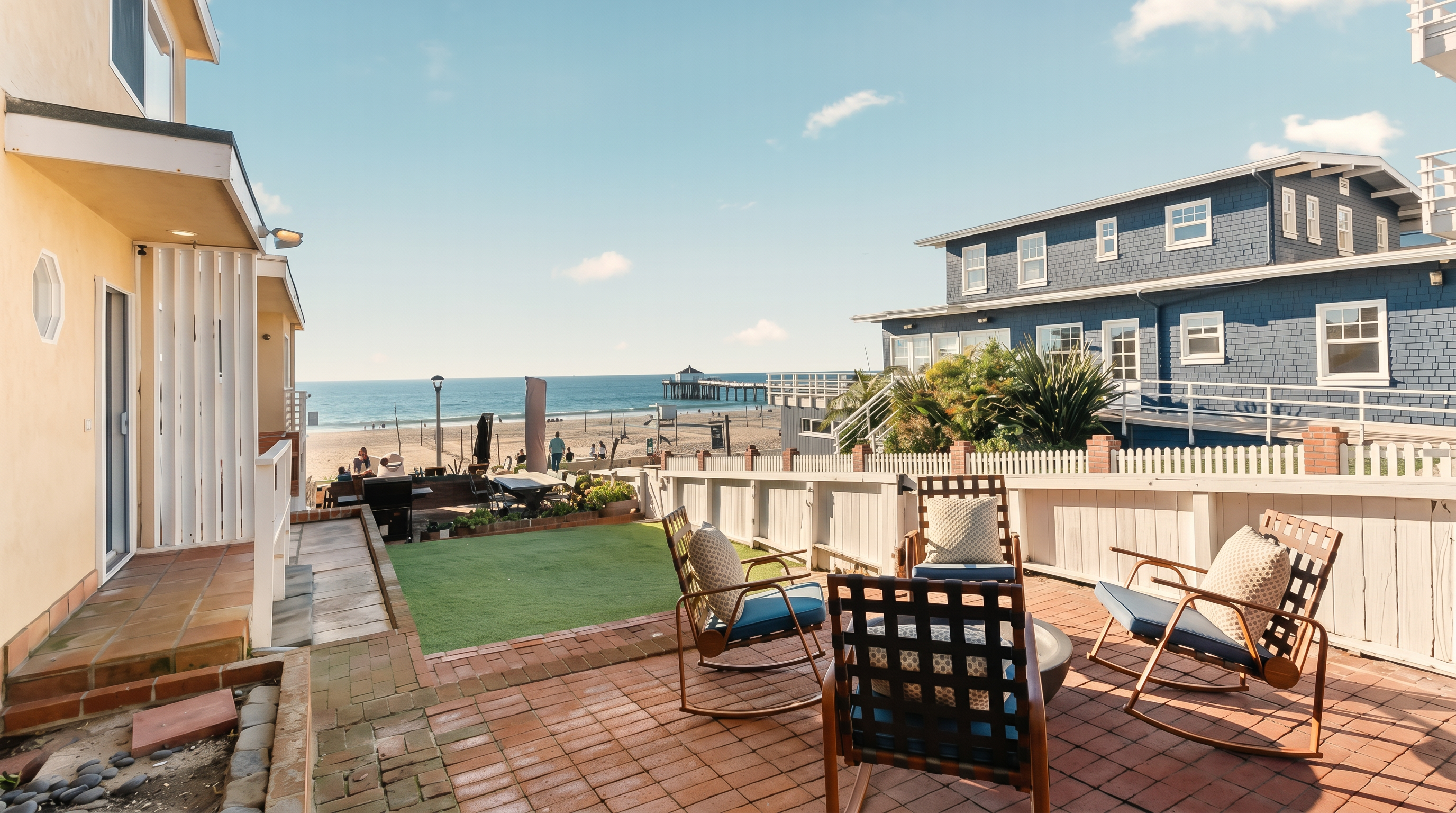 This outdoor living space features a brick-paved patio furnished with several rocking chairs, overlooking a small patch of green lawn and the ocean beyond. A white wooden fence separates the property from the beach, providing a clear view of the nearby pier and coastline. The scene is captured from a ground-level perspective, emphasizing the seamless transition from the private patio to the coastal environment.