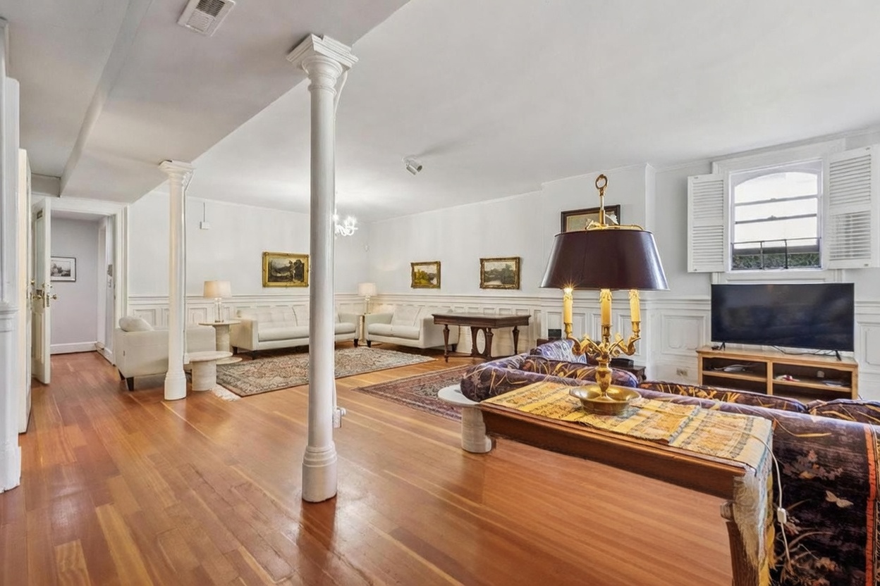 This spacious living area features warm-toned wood flooring and classic architectural details, including white wainscoting and prominent structural columns. The room is furnished with multiple seating areas, including a patterned sofa in the foreground and light-colored sofas in the background, all arranged around area rugs. A large television sits on a wooden media console near a window with open shutters, creating a bright and inviting atmosphere.