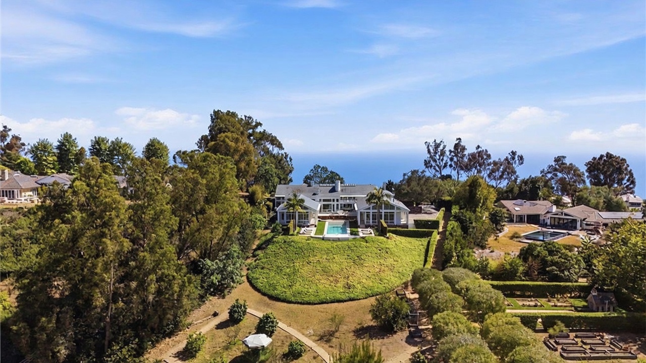 This expansive aerial view captures a sprawling estate perched on a hillside overlooking the ocean. The property features a large residence with a central swimming pool, surrounded by manicured lawns, lush landscaping, and mature trees. The perspective highlights the home's secluded location and its integration into the natural coastal terrain.