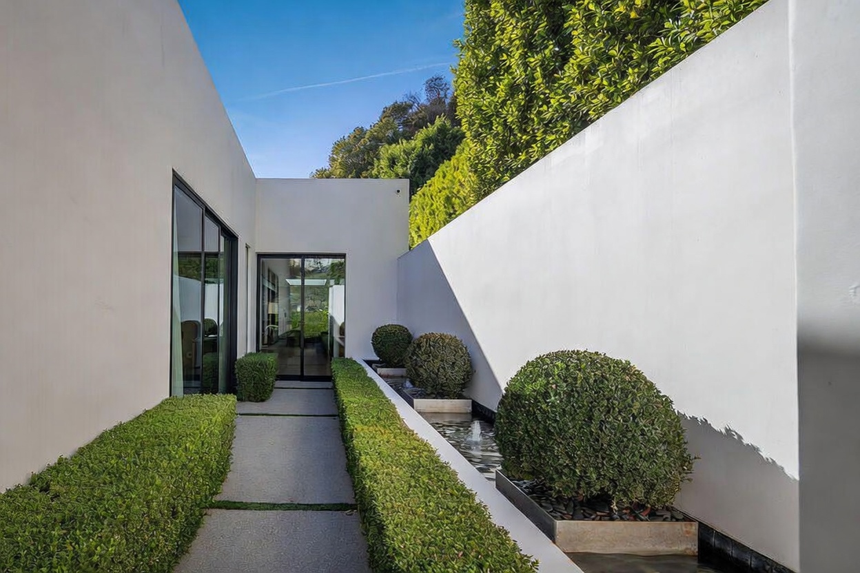 This modern entryway features a minimalist design with crisp white walls and a clean-lined walkway leading to a glass-paneled door. A narrow water feature runs alongside the path, accented by manicured spherical shrubs and a long, low hedge. The bright, sunny atmosphere and geometric landscaping create a sophisticated and serene arrival experience.