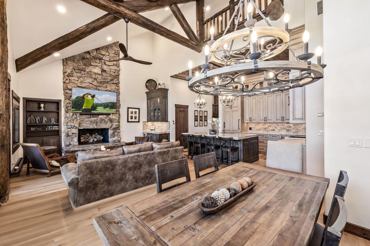 This open-concept living and dining area features a rustic aesthetic with exposed timber beams and a prominent stone-clad chimney breast housing a television above the fireplace. A large, circular chandelier serves as a focal point above the kitchen island, which is surrounded by bar seating and complemented by neutral cabinetry. The space is anchored by a plush, leather-toned sofa and a substantial wooden dining table, creating a warm and inviting atmosphere suitable for entertaining.