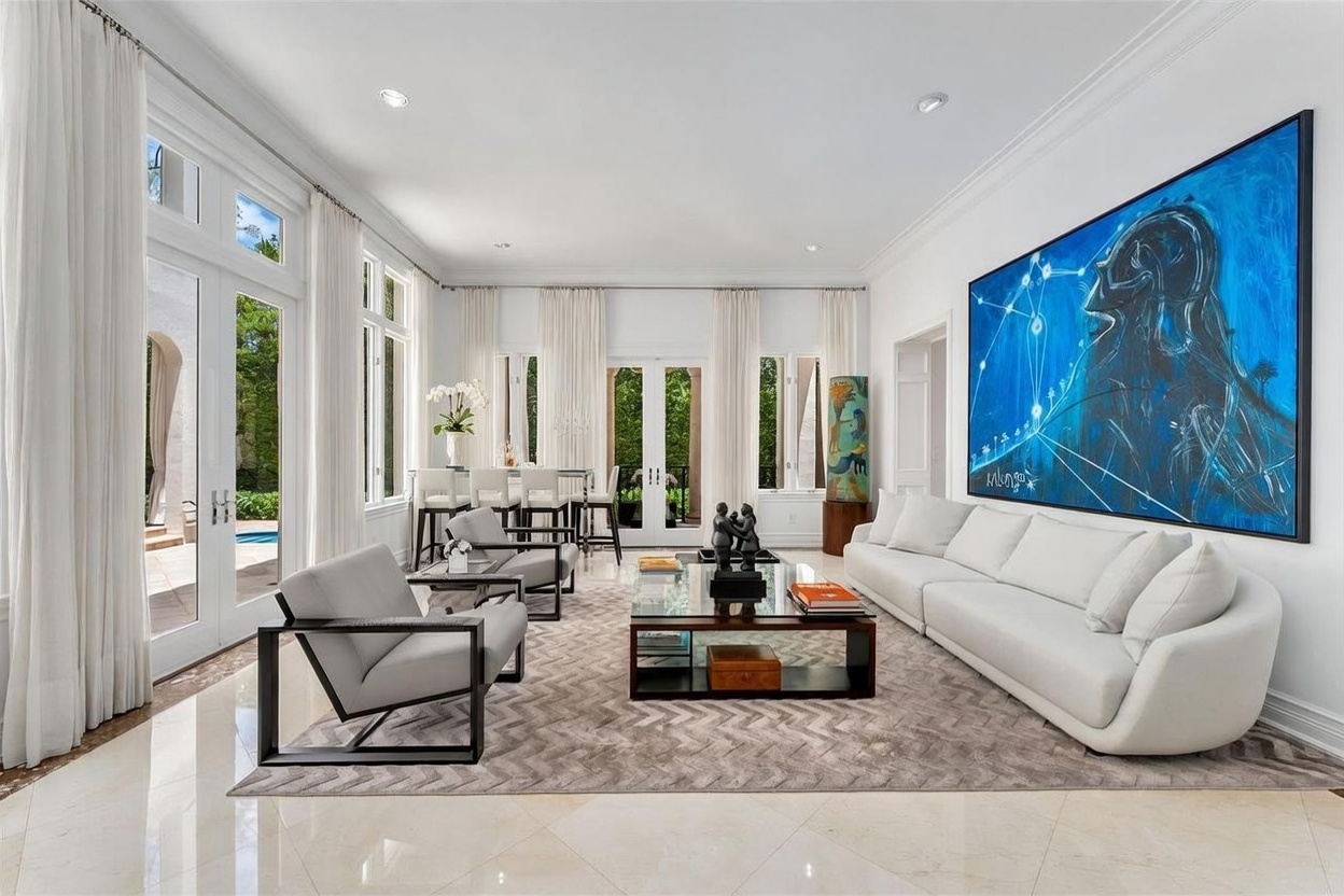 This bright and airy living room features a large white sofa and two grey armchairs arranged around a glass-topped coffee table on a patterned area rug. The space is defined by tall windows and glass doors that provide abundant natural light and views of the outdoor pool area. A prominent, vibrant blue abstract painting serves as a focal point on the wall, while the neutral color palette and polished light-colored flooring create a sophisticated, contemporary atmosphere.