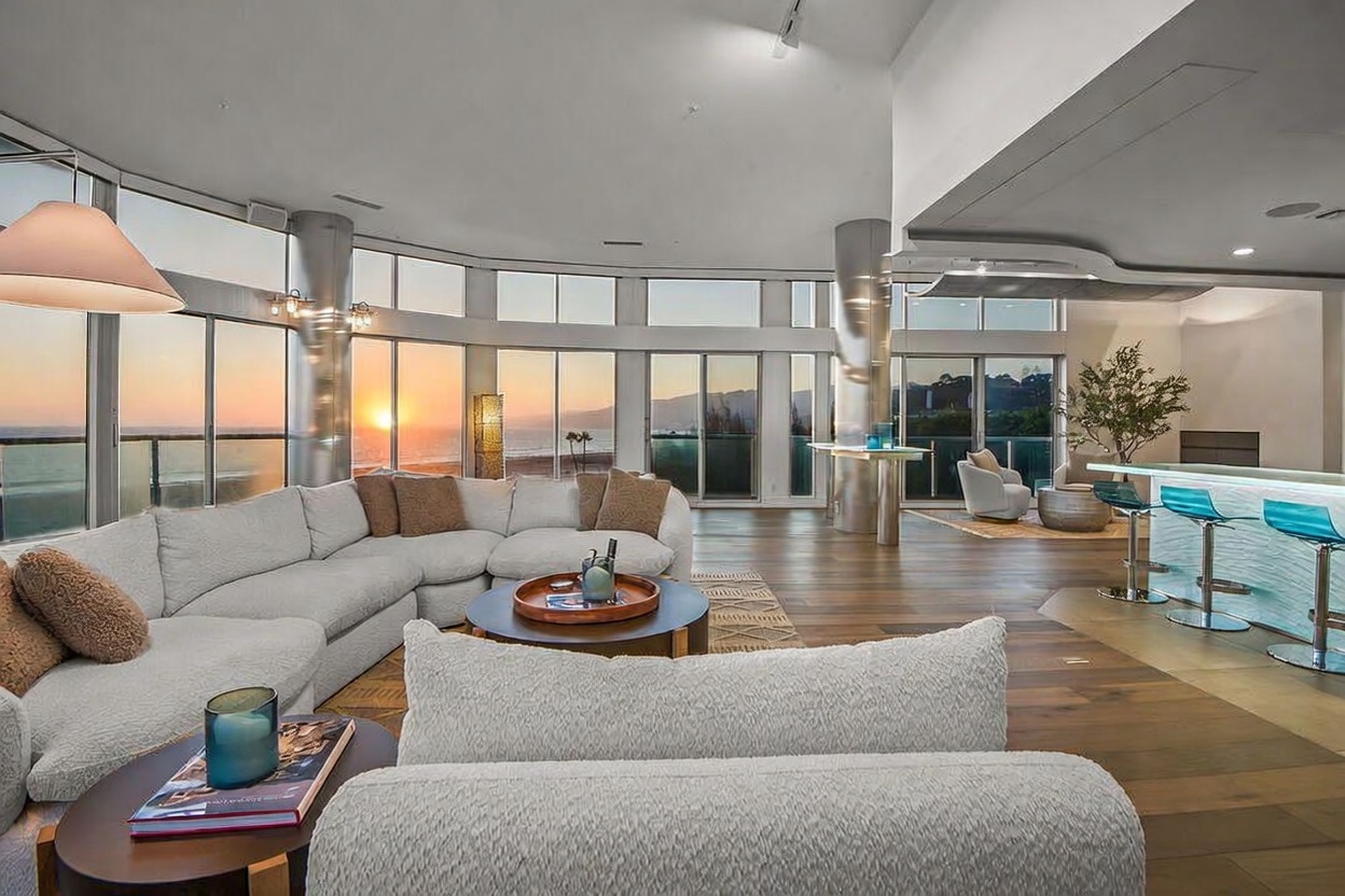 This expansive, open-concept living area features a large, plush sectional sofa arranged around a circular coffee table, set against a backdrop of floor-to-ceiling windows that offer a panoramic sunset view of the ocean. The space includes a sleek bar area with high stools and polished wood flooring, creating a sophisticated and airy atmosphere. The perspective is from the center of the room, looking out toward the horizon, emphasizing the seamless connection between the interior living space and the coastal scenery.