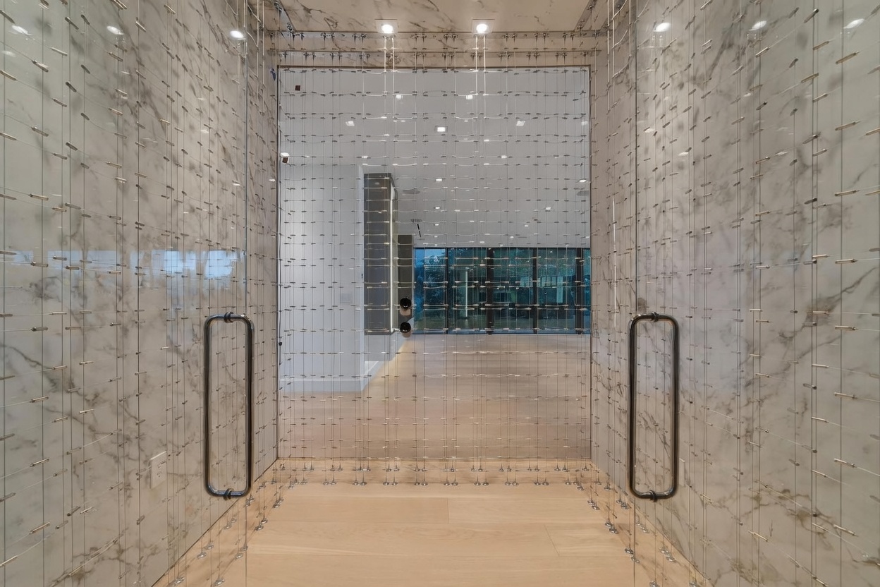 This sophisticated wine cellar features a unique metal cable display system mounted against walls clad in light-toned, veined stone. The space is accessed through a glass door with a prominent dark metal handle, creating a sleek and modern aesthetic. The perspective looks directly through the glass partition into an open, bright living area beyond, emphasizing the home's open-concept design.