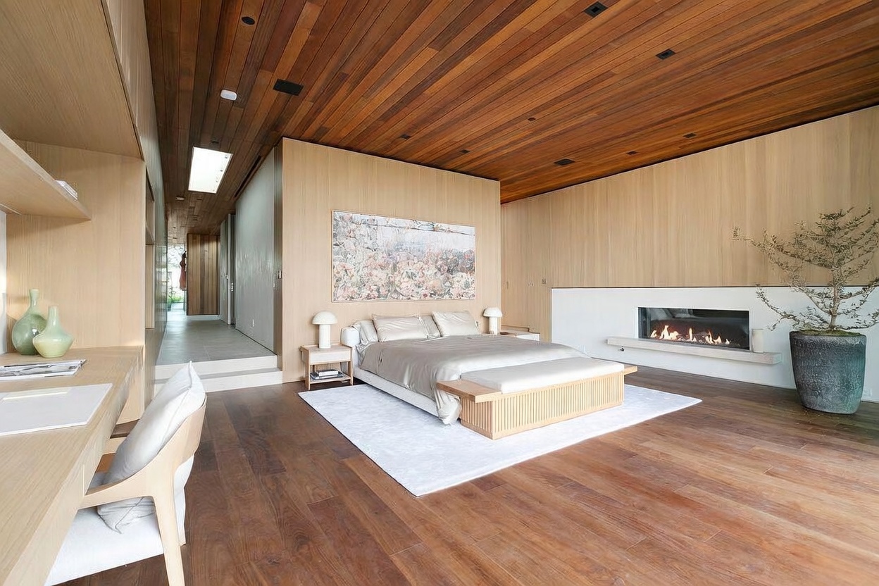 This luxurious primary bedroom features a warm, minimalist aesthetic with rich wood-paneled ceilings and light oak wall accents. The room is anchored by a platform bed with a neutral-toned bedding set, a sleek linear fireplace, and a dedicated workspace area to the left. A large, abstract piece of art hangs above the bed, while a potted plant adds a touch of organic texture to the serene, high-end space.