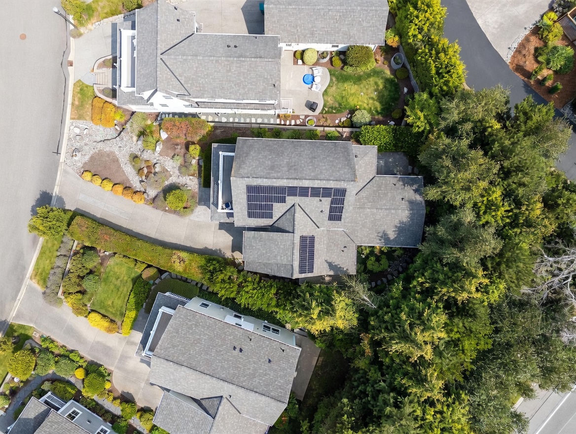 This high-angle aerial view captures a residential property nestled within a lush, tree-lined neighborhood. The home features a complex shingled roof equipped with solar panels, surrounded by manicured landscaping, paved driveways, and mature greenery. The perspective provides a clear layout of the house's footprint and its integration into the surrounding suburban environment.